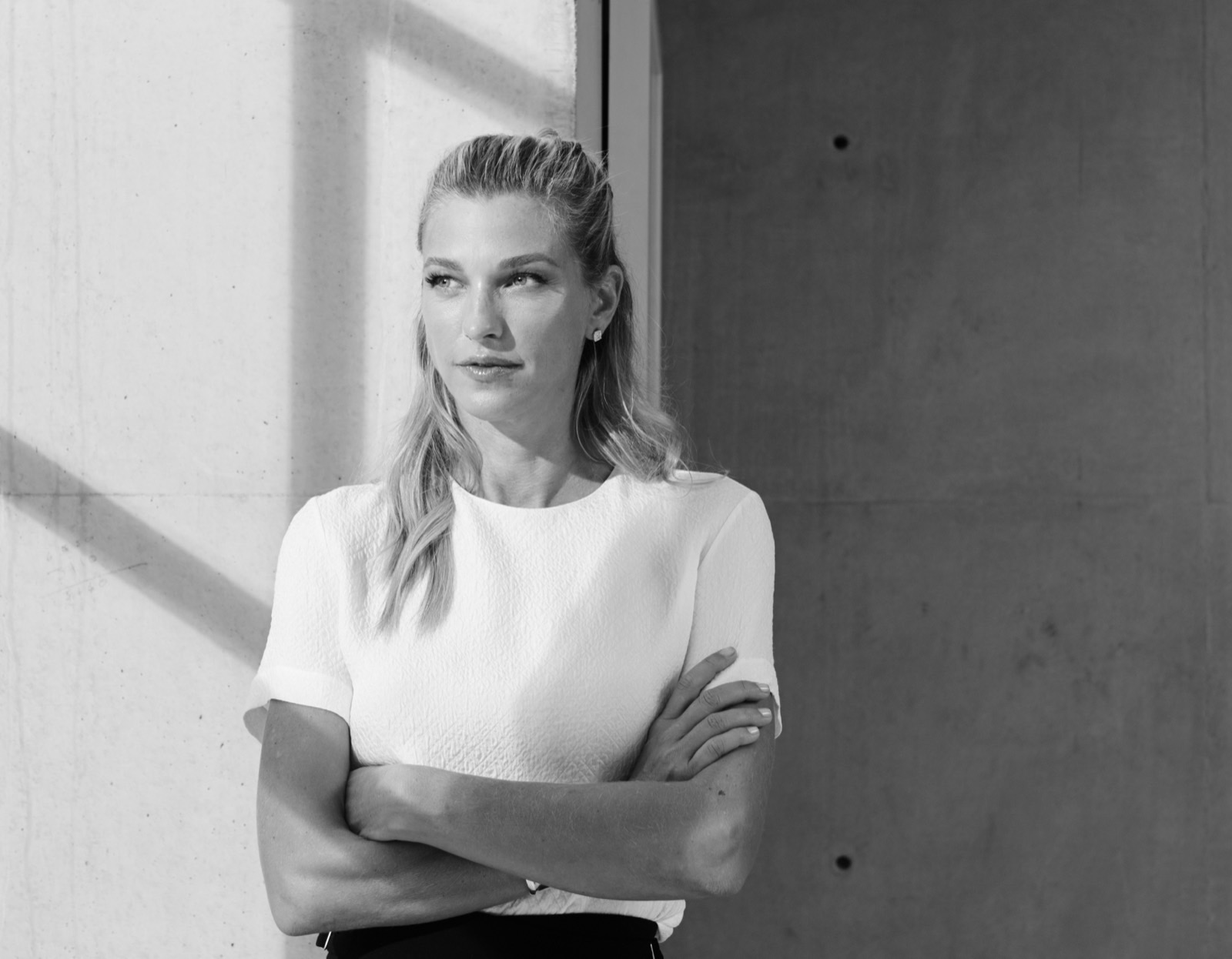 Nicole Junkermann in an ivory blazer with her arms crossed against a pale concrete wall, a sleek modern portrait